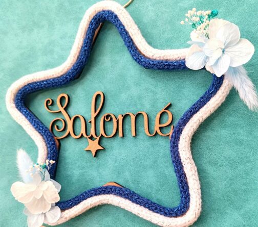 Une étoile en bois avec des fleurs blanches et bleues et le prénom salomé au centre . Le prénom est entouré de deux tricotins en forme d’étoile , un tricotin bleu et blanc brillant.